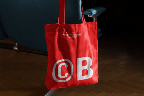 Red tote bag on a blue chair with bold C B logo design. Ideal for branding mockups and designer presentations. Modern design accessory asset.