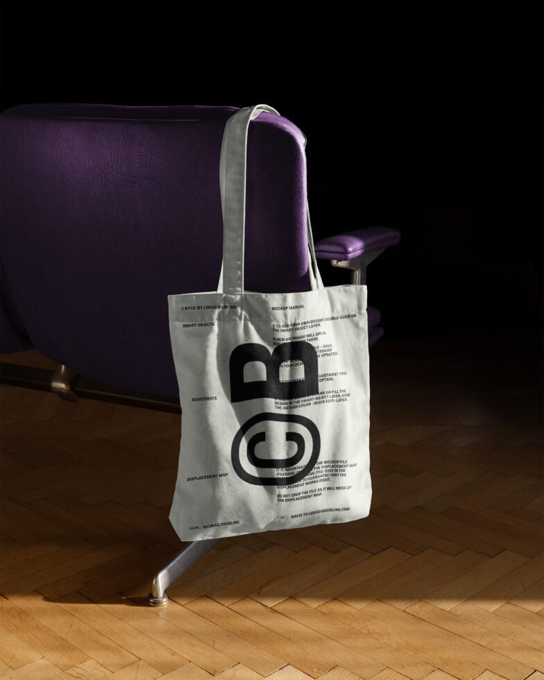 Tote bag mockup with bold letters displayed on a wooden floor beside a purple chair. Perfect for showcasing branding or logo designs for designers.