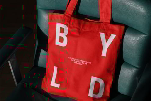 Red tote bag with bold white letters BYLD on green chair. Ideal for branding mockup. Design for creatives, showcasing typography and layout skills.