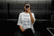 Person in elevator wearing a blank white T-shirt with Mockup Lab text holding phone. Fashion template for designers. SEO Keywords: Mockup, T-shirt, Template.