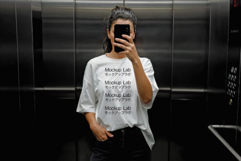 Person in elevator wearing a blank white T-shirt with Mockup Lab text holding phone. Fashion template for designers. SEO Keywords: Mockup, T-shirt, Template.