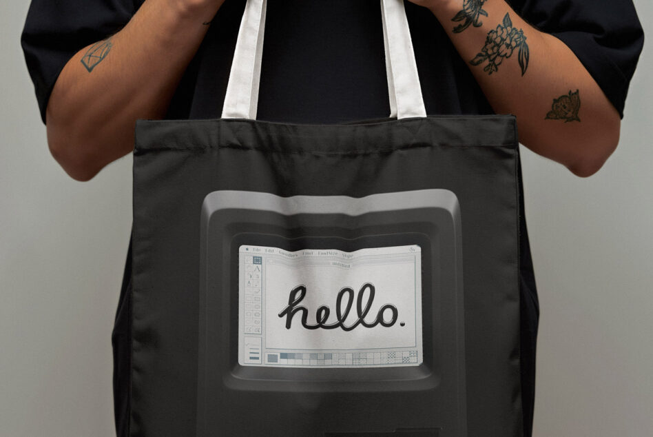 Black tote bag with retro computer monitor showing hello text worn by person with tattoos