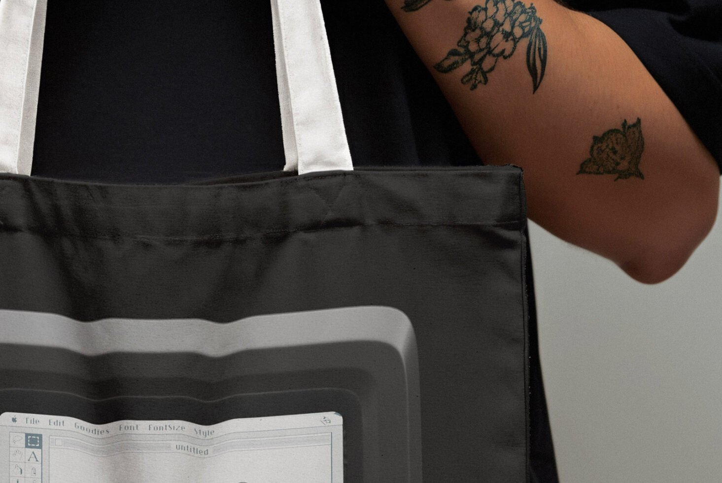 Tote bag with nostalgic computer interface design in grayscale held by tattooed person. Ideal for graphic designers seeking unique printable templates or mockups.