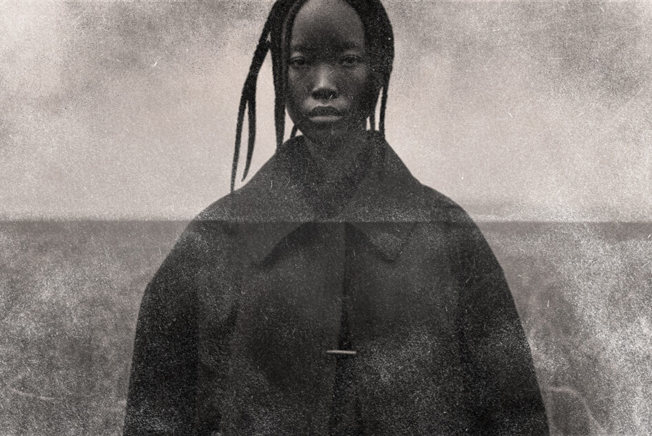 Grainy black portrait of figure with braids facing camera by ocean horizon