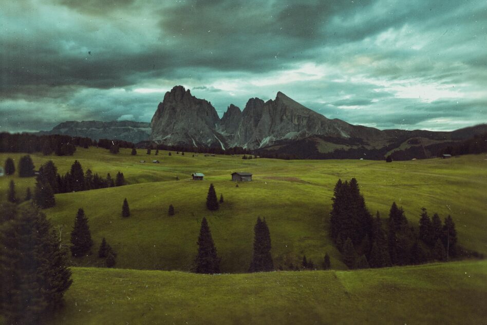 Alpine valley with dramatic peaks, scattered cabins, pine forest, moody sky