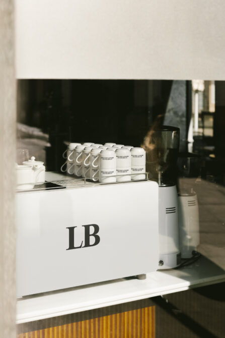 Coffee shop window showing sleek white coffee machine with LB logo and stacked cups. Ideal for branding mockups or coffee shop design templates.