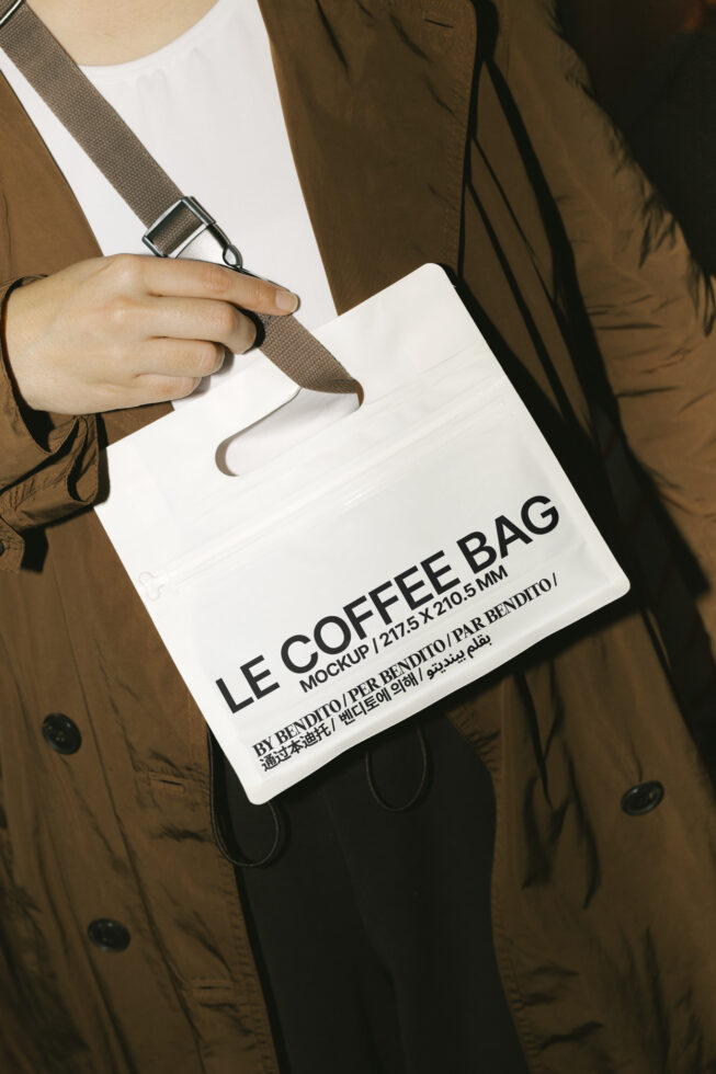 Person holding a white coffee bag mockup with text in multiple languages, brown strap. Ideal for branding and packaging design. Mockups, Designers, Templates.