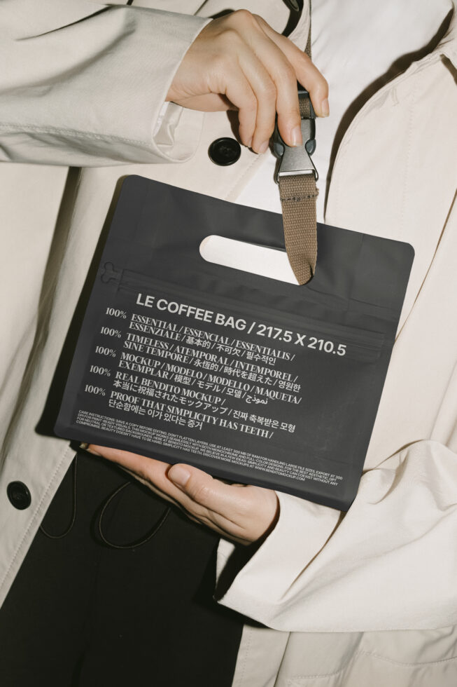 Person holding black coffee bag mockup with text in multiple languages. Ideal for graphic design, packaging templates, and designer resources.