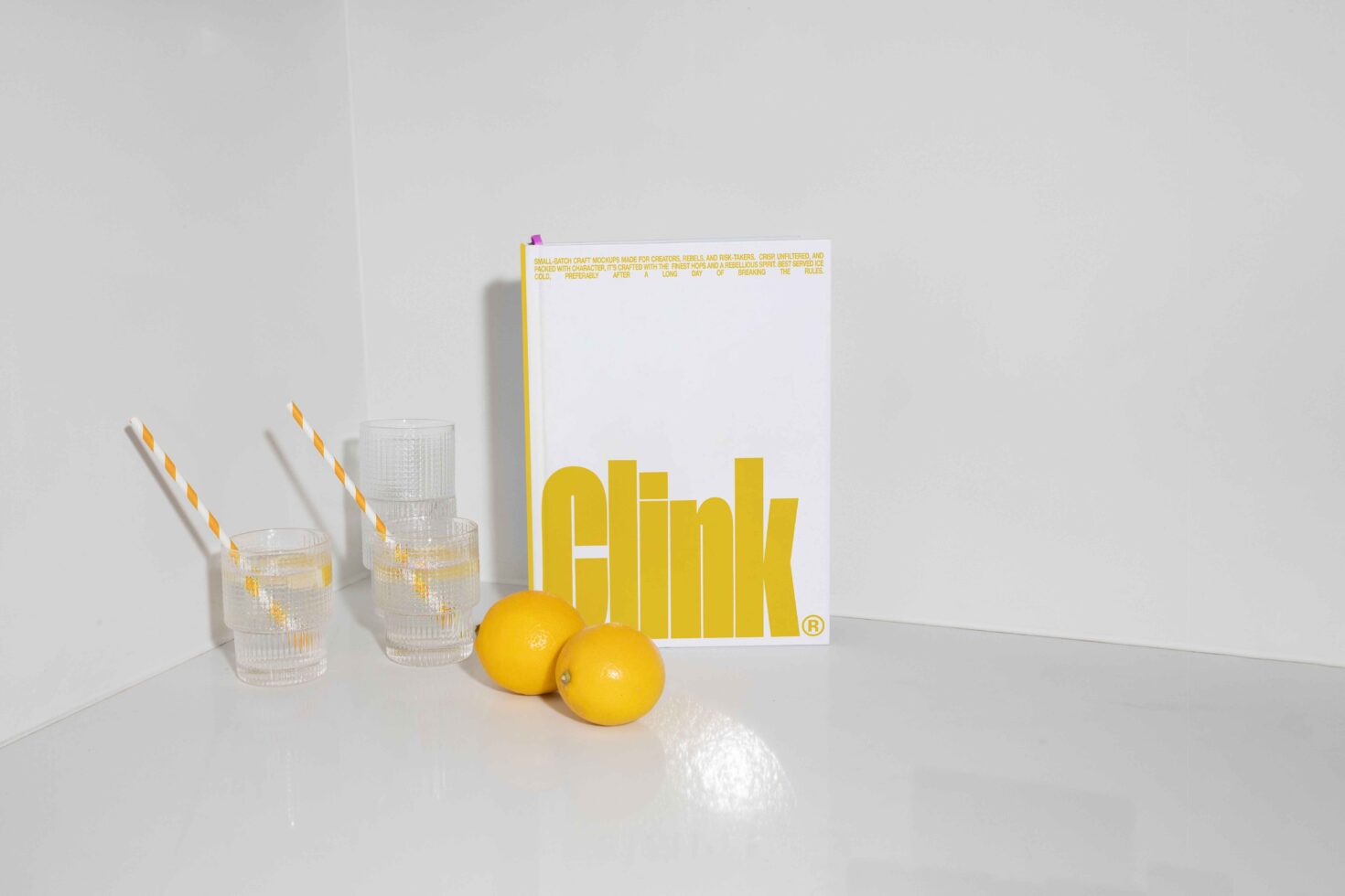 Minimalist mockup with a white book featuring bold yellow text Clink. Three glass cups with striped straws and two lemons on a white surface.
