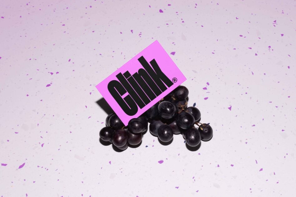 Hot pink card with black text propped on dark grapes over lavender surface