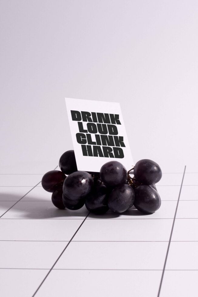 Black grape bunch with a card displaying bold text Drink Loud Clink Hard on a minimalist grid background suitable for mockups and graphic design.