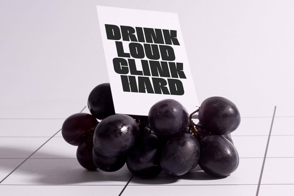 White card in front of dark grapes displaying black chunky letterforms on light backdrop