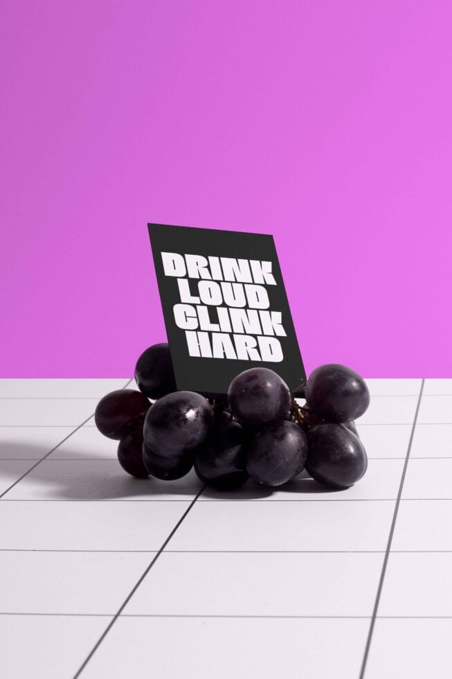 Black card with bold text Drink Loud Clink Hard on grapes over checkered surface against pink background mockup graphic design template vibrant modern