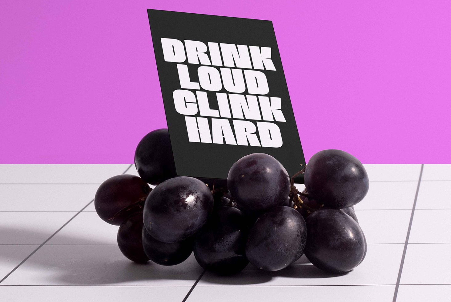 Black card with bold white font text Drink Loud Clink Hard sits on grapes on a tiled surface against a purple background. Font design mockup.