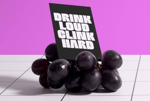 Black card with bold white font text Drink Loud Clink Hard sits on grapes on a tiled surface against a purple background. Font design mockup.