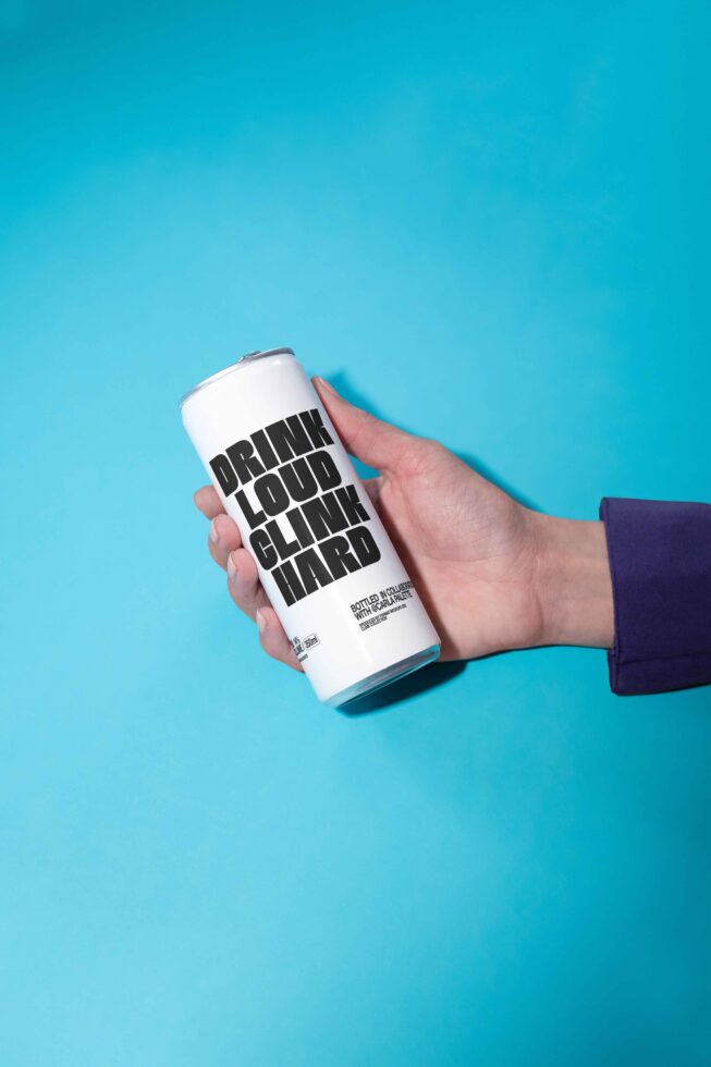 A hand holds a white can with bold black text Drink Loud Clink Hard on a blue background. Ideal for mockups fonts packaging design templates.