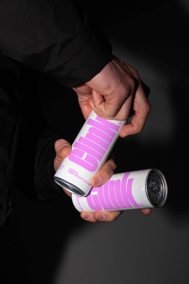 Person holding modern beverage cans with bold pink typography design against a dark background mockup template for designers branding packaging.
