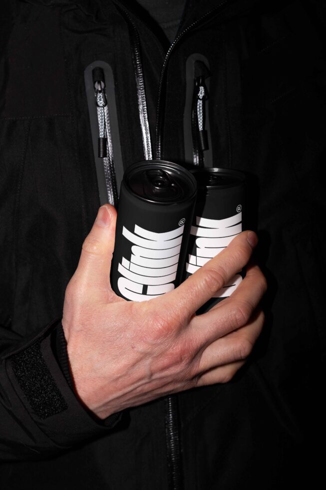 Person holding two sleek black cans with bold white branding, wearing a dark jacket. Ideal for product mockups, branding design, packaging templates.