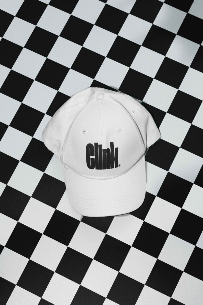 White cap with bold black text design placed on a black and white checkerboard background. Perfect for apparel mockups and graphic design projects.