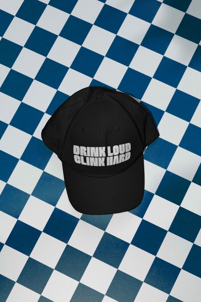Black cap with bold white text Drink Loud Clink Hard on a blue and white checkered background suitable for mockup use by graphic designers