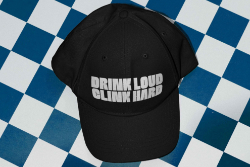 Black baseball cap mockup with bold white text Drink Loud Clink Hard on a blue and white checkered surface. Ideal for showcasing custom designs