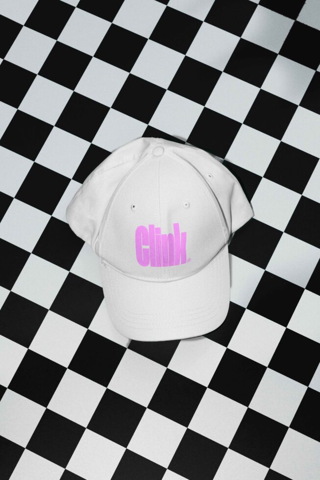 White baseball cap with pink text sits on black-and-white checkered surface. Ideal for mockup projects, design templates, fashion graphics, and branding.