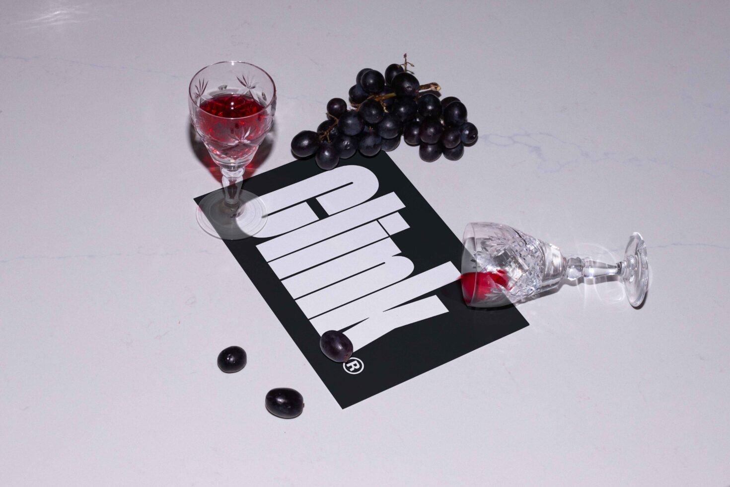 Wine glasses with red liquid, grape bunches and bold text on a card lay on white surface. Ideal for mockup, branding, product design visuals.