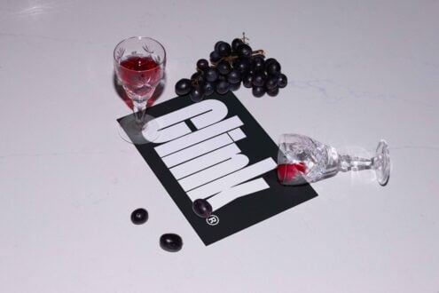 Wine glasses with red liquid, grape bunches and bold text on a card lay on white surface. Ideal for mockup, branding, product design visuals.