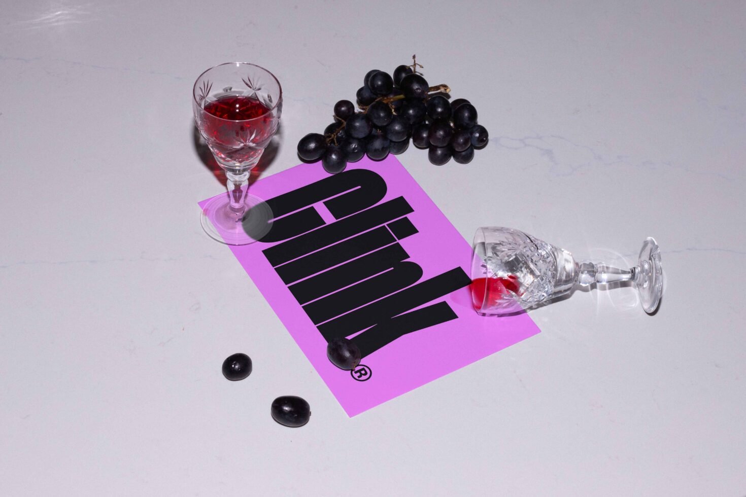 Wine glasses with grapes on a surface featuring bold typography design on pink paper ideal for graphic templates and design mockups.