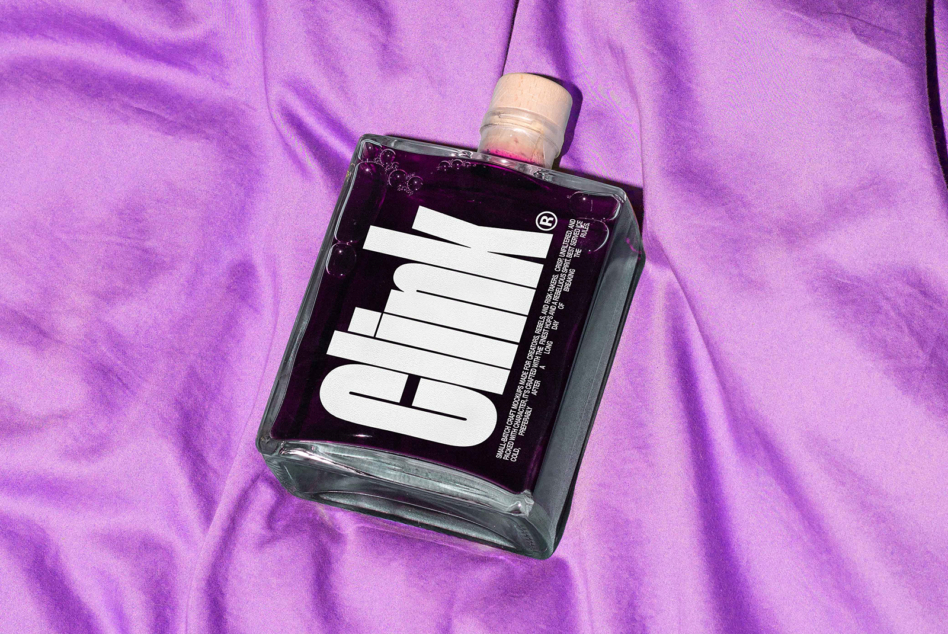Glass bottle with bold typography label on purple fabric background. Ideal for mockups and graphic design projects. High-quality designer asset.
