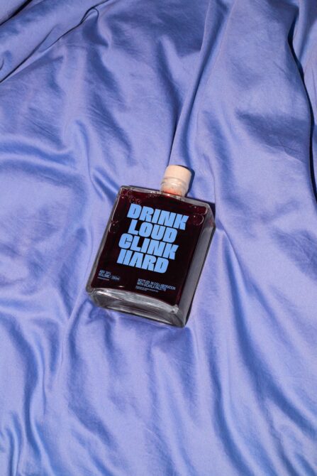 Square bottle with blue label reading Drink Loud Clink Hard, resting on a blue fabric background. Keywords: mockup, bottle design, packaging.