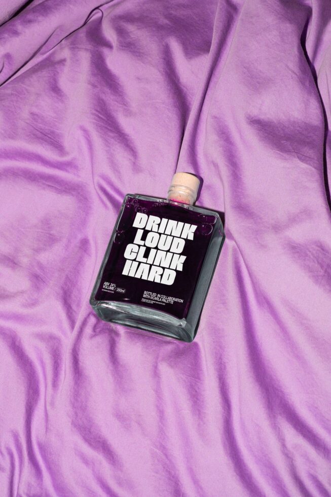 Purple liquid-filled bottle with Drink Loud Clink Hard label on crumpled lavender fabric background ideal for graphic design or branding templates.