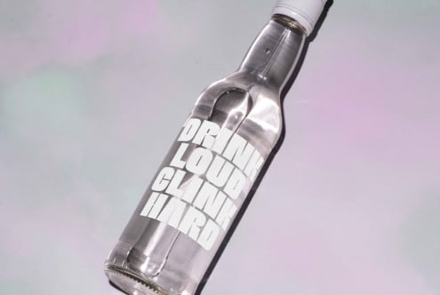 Clear glass bottle with bold typography labels Drink Loud Clink Hard on soft gradient backdrop ideal for designers mockup projects branding display.