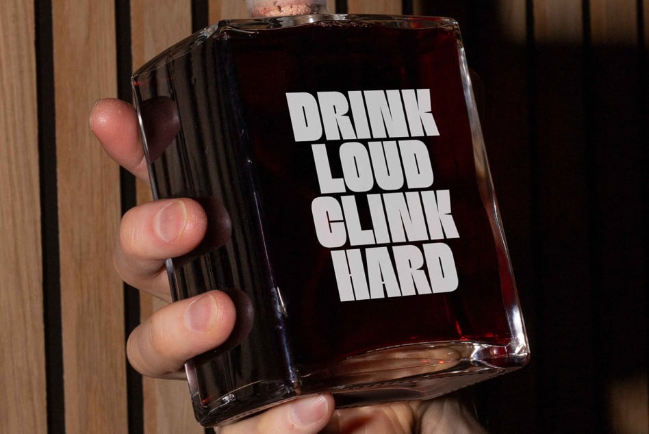 Person holding bottle with Drink Loud Clink Hard text in white on dark wood backdrop