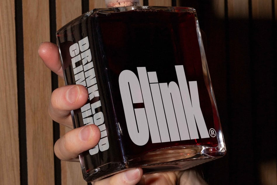 Hand gripping square bottle with white Olink branding on dark liquid, wooden slat background