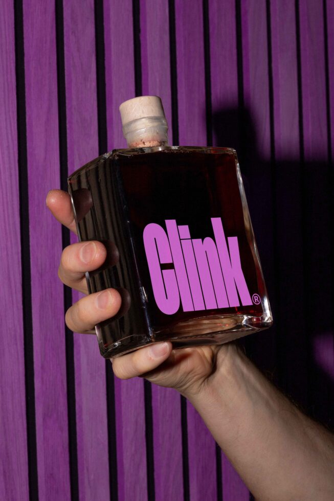 Square glass bottle with bold pink text logo Clink held against a purple striped wall. Ideal mockup for designers focused on branding and packaging visuals.