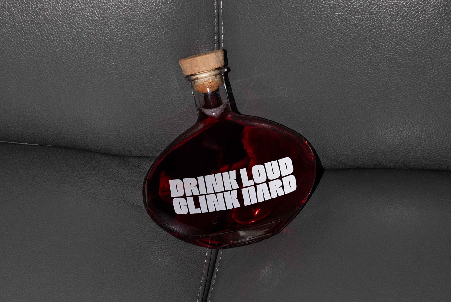 Oval glass bottle with dark liquid and wooden cap on grey leather. Text Drink Loud Clink Hard. Ideal for branding mockups, graphic designers, template assets.
