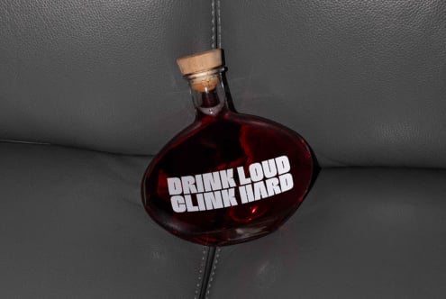 Oval glass bottle with dark liquid and wooden cap on grey leather. Text Drink Loud Clink Hard. Ideal for branding mockups, graphic designers, template assets.