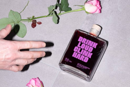 Flat lay of a glass bottle with bold typography reading Drink Loud Clink Hard next to a hand and roses on a tile surface mockup design elements