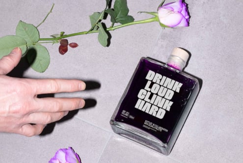 Hand reaching for a purple-colored bottle labeled Drink Loud Clink Hard beside a rose on a tiled surface. Perfect for mockups and design assets.