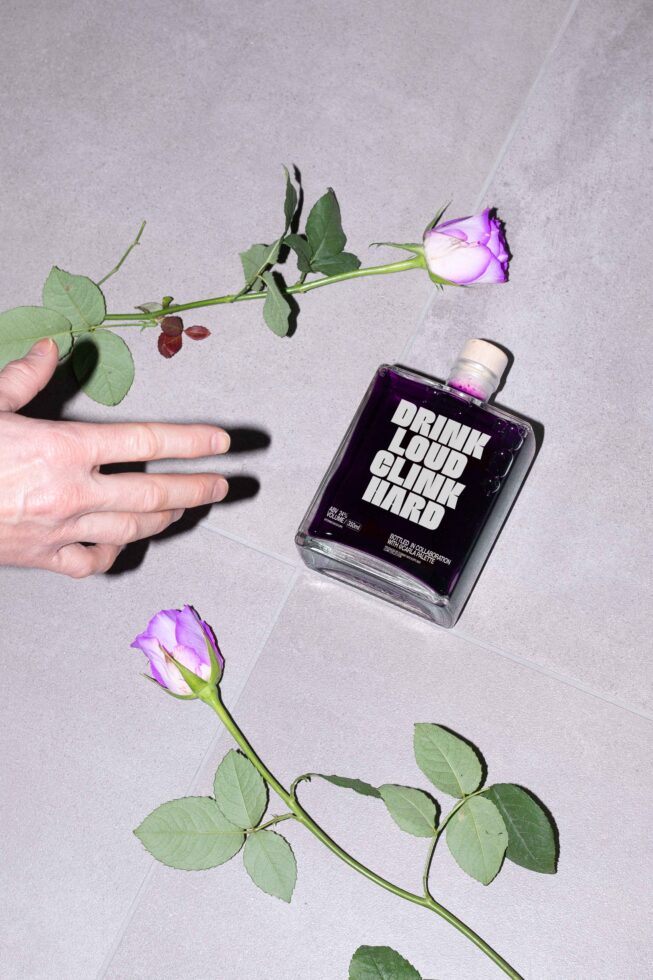 Bottle with bold text Drink Loud Clink Hard on gray surface next to two purple roses and hand. Perfect for product mockup or design template use.
