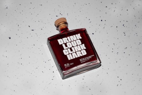 Glass bottle with bold typography Drink Loud Clink Hard on speckled surface. Ideal for designers seeking graphics or packaging mockups.