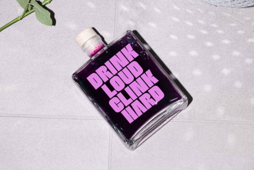 Glass bottle with purple liquid on a tiled surface, bold pink text Drink Loud Clink Hard, suitable for graphic design mockup, packaging design.