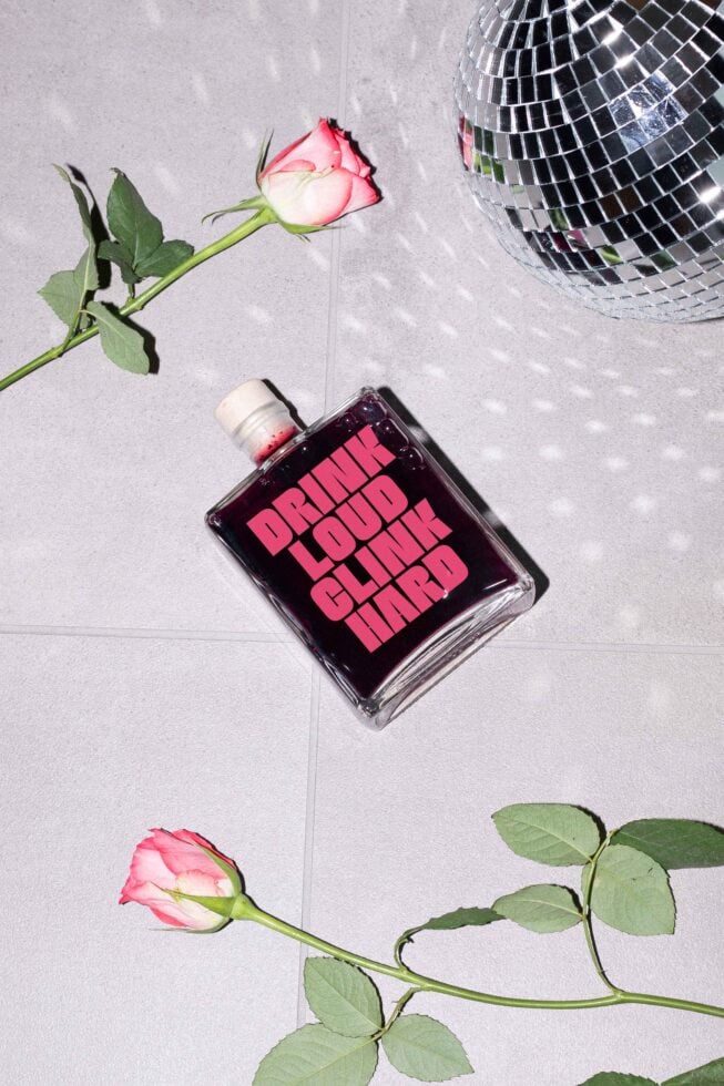 Flat lay of a square glass bottle with bold typography Drink Loud Clink Hard. Two pink roses and disco ball on tiled surface. Party theme mockup.