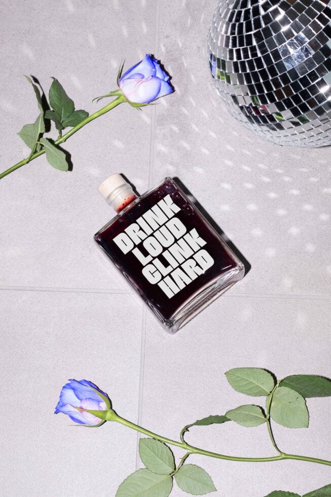 Flat lay design featuring a bottle with bold text Drink Loud Clink Hard a disco ball reflecting light and blue roses ideal for trendy graphic templates
