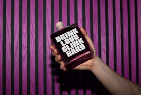 Hand holding a bottle with bold white text Drink Loud Clink Hard against a striped purple and black background ideal for designers digital mockup asset.