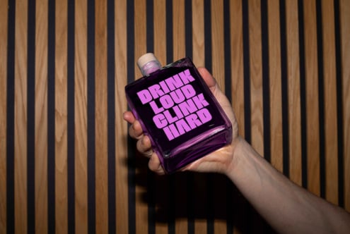 Hand holds a purple bottle with bold text Drink Loud Clink Hard against a striped wooden background. Perfect for mockups or branding templates.