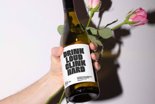Hand holding a wine bottle with bold typography on label Drink Loud Clink Hard paired with pink roses ideal for graphic design mockups branding projects
