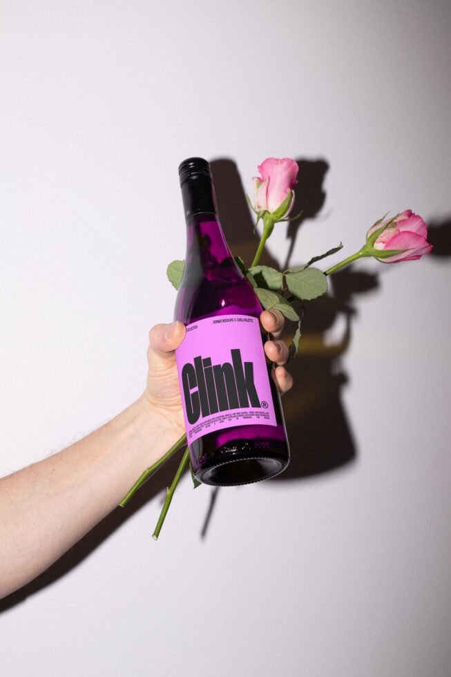 Hand holding a bottle with bold typography label next to pink roses on white background perfect for mockups branding packaging design templates.