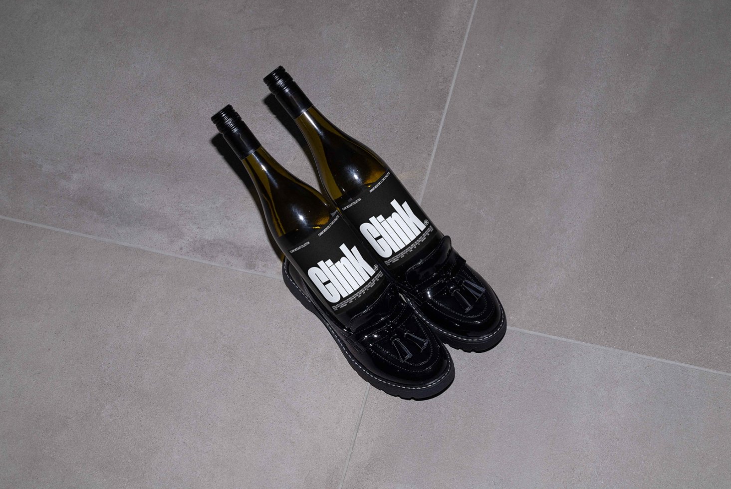 Two wine bottles creatively set as shoes with black glossy loafer shoes on gray tiled floor. Clink branding visible. Unusual product design mockup.